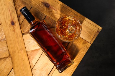 Whiskey with ice cubes in glass and bottle on wooden crate against black background, top view