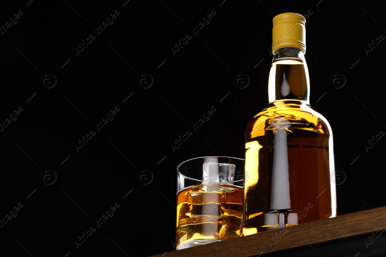 Photo of Whiskey with ice cubes in glass and bottle on wooden table against black background, low angle view. Space for text