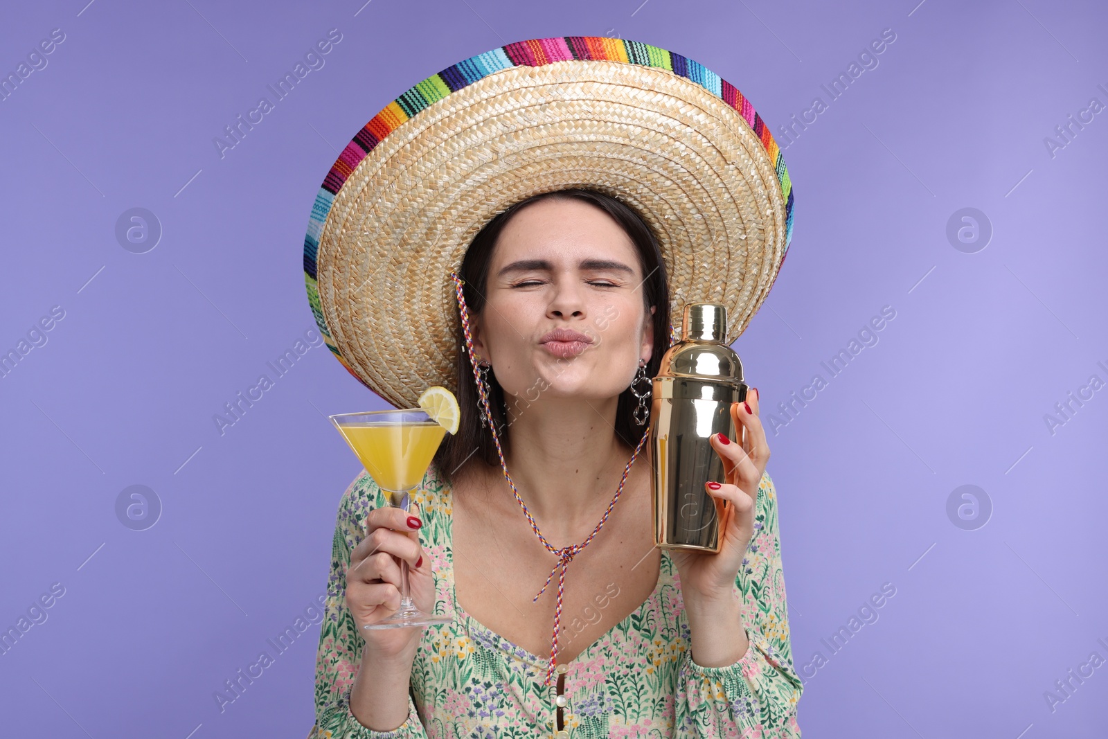 Photo of Young woman in Mexican sombrero hat with cocktail and shaker sending air kiss on violet background