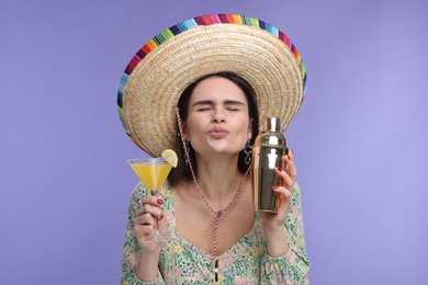 Young woman in Mexican sombrero hat with cocktail and shaker sending air kiss on violet background