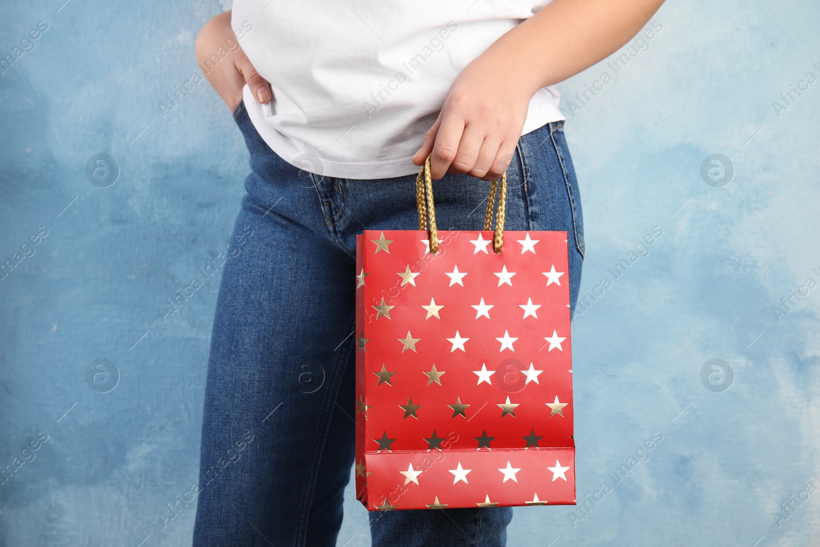 Photo of Woman holding red shopping paper bag with star pattern on light blue background, closeup