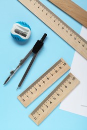 Different rulers, pencil sharpener and compass on light blue background, flat lay