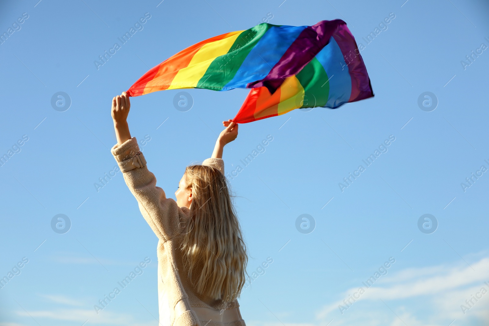 Photo of Woman holding bright LGBT flag against blue sky