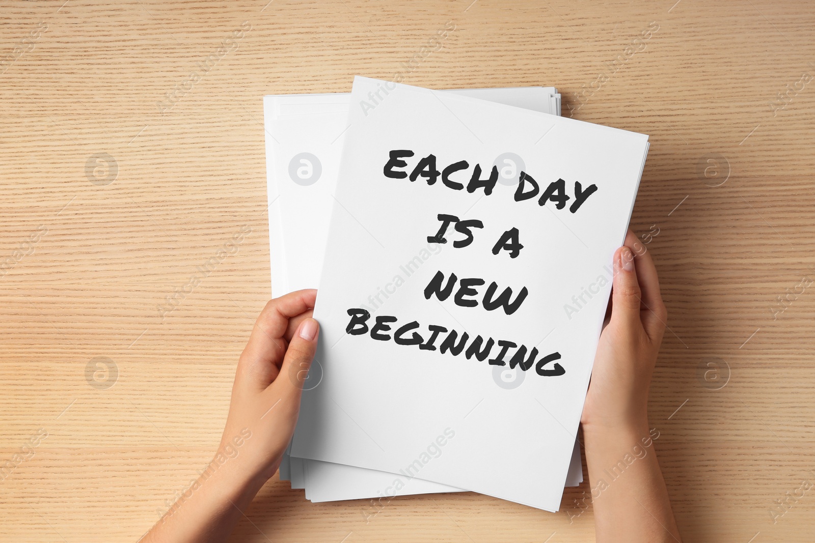 Image of Woman holding paper with inspirational text Each Day Is A New Beginning at wooden table, top view. Change your life