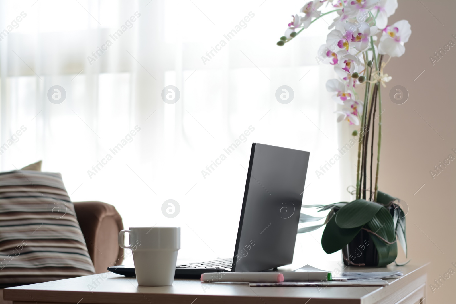 Photo of Ceramic cup, laptop and stationery near blooming orchid on wooden table indoors. Good morning