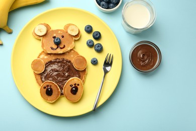 Photo of Creative serving for kids. Plate with cute bear made of pancakes, blueberries, bananas and chocolate paste on light blue table, flat lay