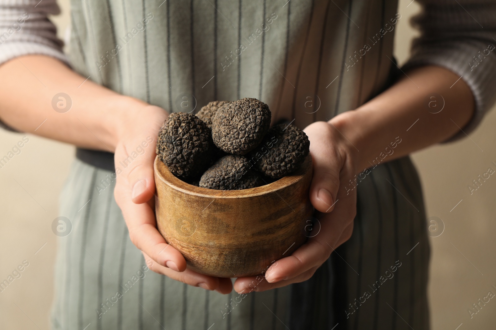 Photo of Woman holding wooden bowl of black truffles in hands on beige background, closeup