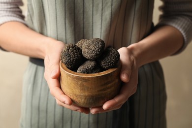 Photo of Woman holding wooden bowl of black truffles in hands on beige background, closeup