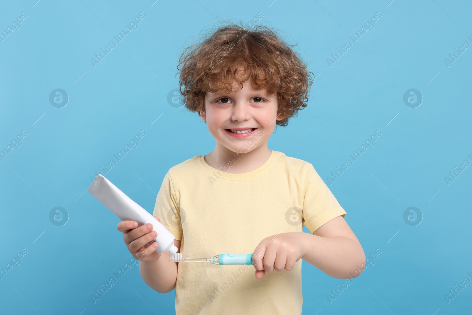 Photo of Cute little boy squeezing toothpaste from tube onto electric toothbrush on light blue background