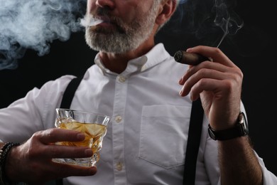 Man with glass of whiskey smoking cigar on black background, closeup