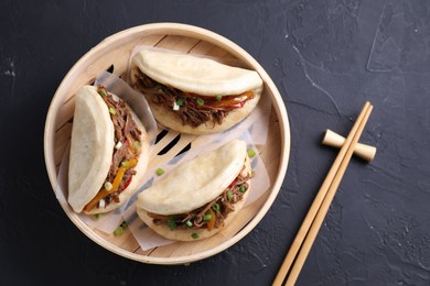 Photo of Delicious gua bao in bamboo steamer and chopsticks on black table, top view