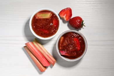 Tasty rhubarb jam in bowls, cut stems and strawberries on white wooden table, flat lay