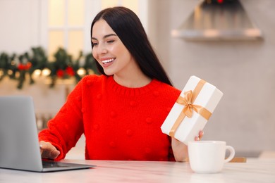 Celebrating Christmas online with exchanged by mail presents. Smiling woman with gift box during video call on laptop at home