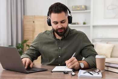 Photo of E-learning. Young man taking notes during online lesson at wooden table indoors