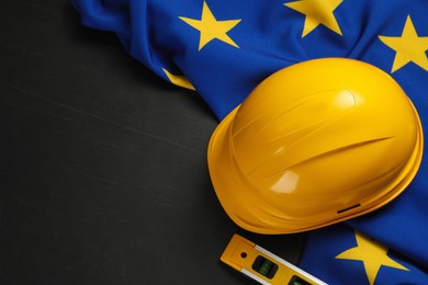 Yellow hard hat, building level and flag of European Union on black table, flat lay. Space for text