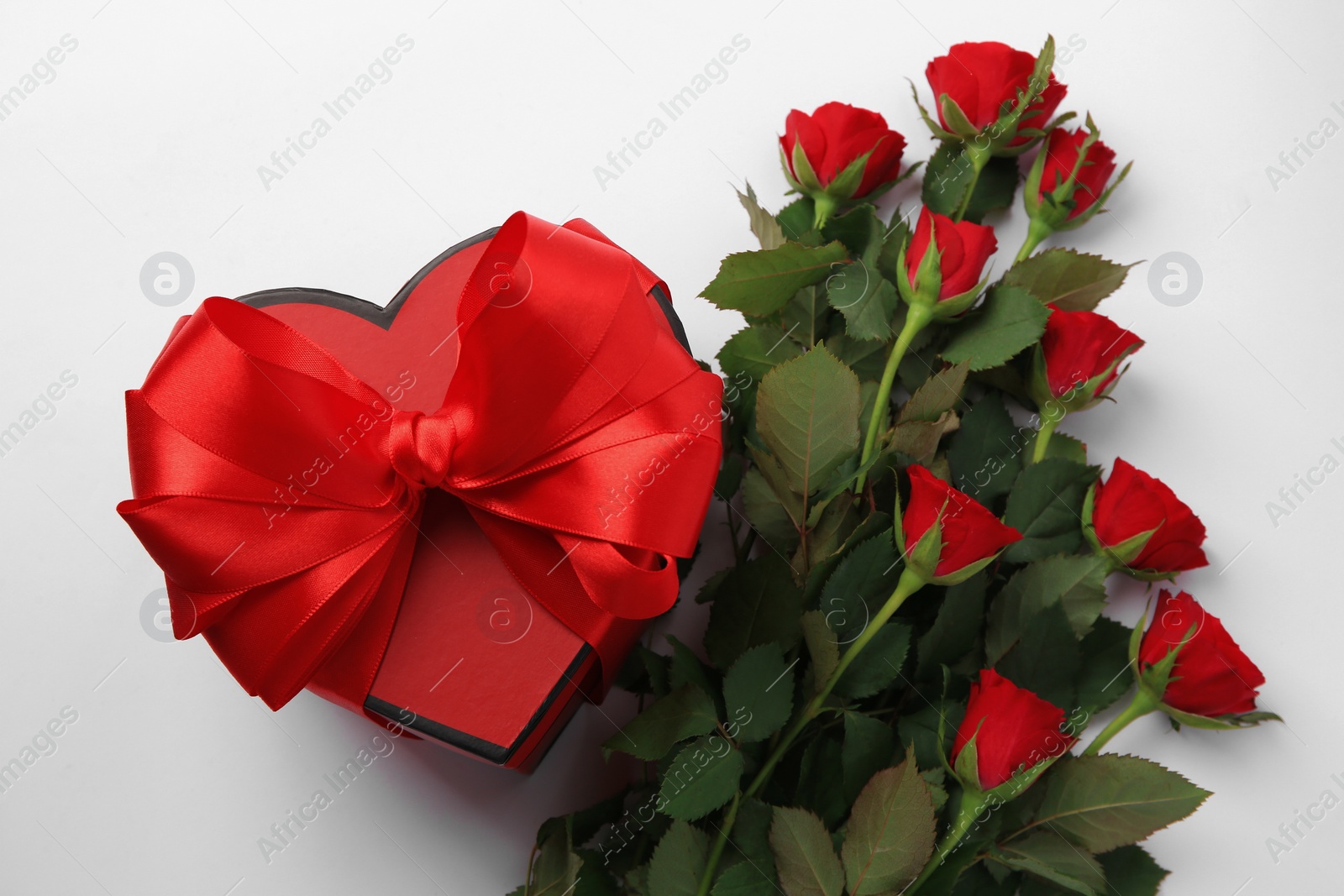 Photo of Heart shaped gift box with bow and beautiful red roses on white background, flat lay