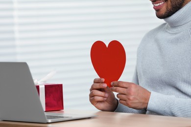 Photo of Valentine's day celebration in long distance relationship. Man holding red paper heart while having video chat with his girlfriend via laptop, closeup