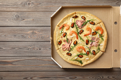 Photo of Delicious seafood pizza in cardboard box on wooden table, top view. Space for text