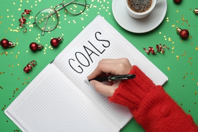 Woman writing in notebook on green background with Christmas decor, above view. New aims