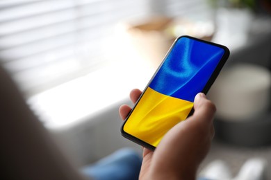Image of Man holding smartphone with Ukrainian national flag on screen indoors, closeup