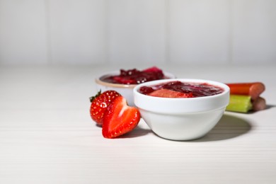 Photo of Tasty rhubarb jam in bowls, stems and strawberries on white wooden table. Space for text