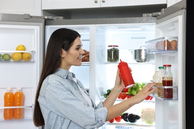 Young woman taking bottle of ketchup out of refrigerator