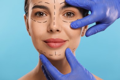 Doctor checking marks on woman's face for cosmetic surgery operation against light blue background, closeup