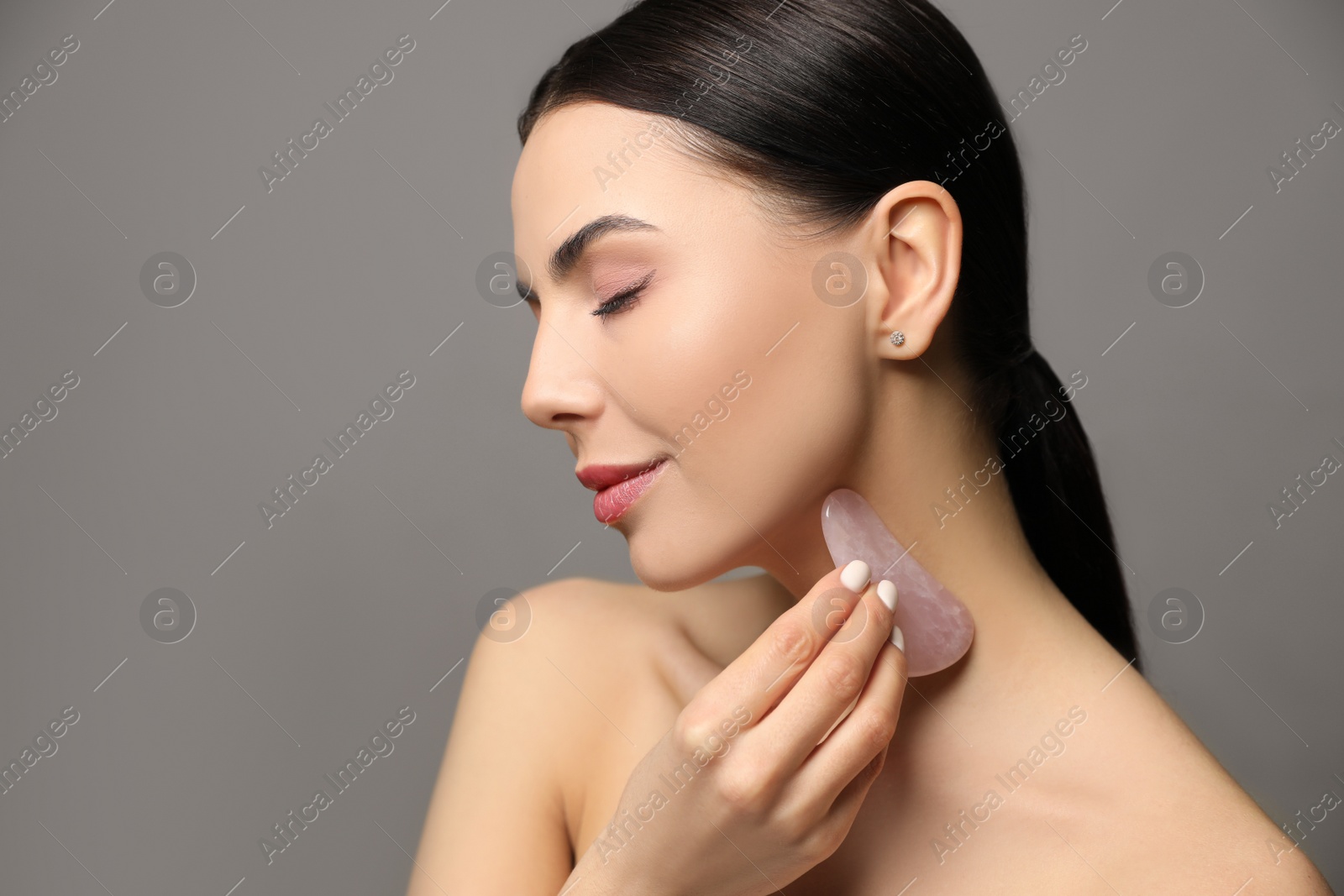 Photo of Beautiful young woman doing facial massage with gua sha tool on grey background, closeup