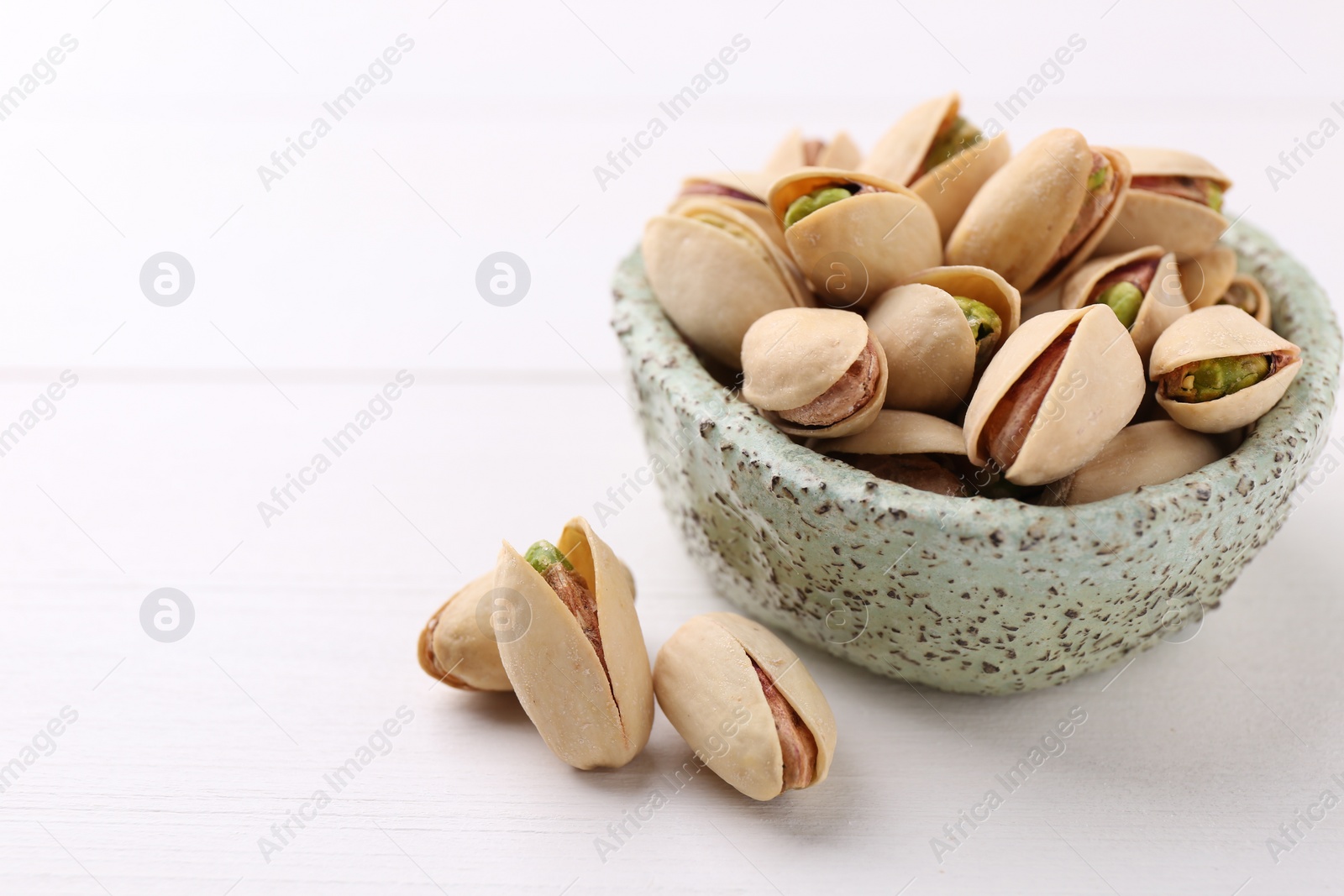 Photo of Delicious pistachios in bowl on white table, closeup. Space for text