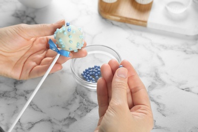 Young woman decorating tasty cake pop with blue sprinkles at white marble table, closeup