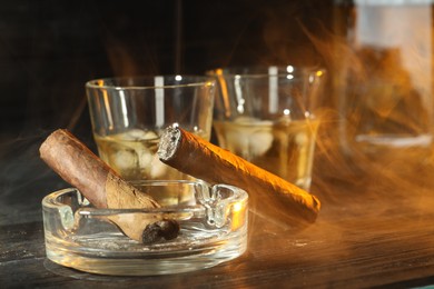 Photo of Cigars, ashtray and whiskey with ice cubes on black wooden table, closeup