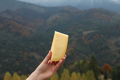 Photo of Woman holding piece of delicious cheese against mountain landscape, closeup