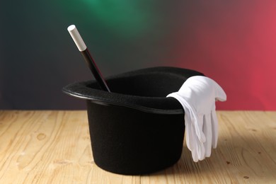 Magician's hat, wand and gloves on wooden table against color background