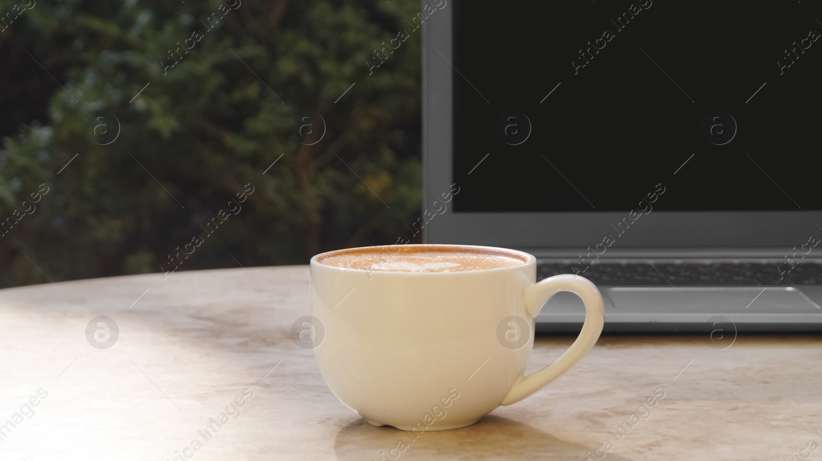 Photo of Cup of delicious coffee and laptop on beige marble table outdoors