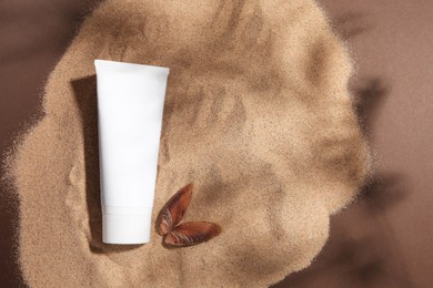 Photo of Tube of body cream and seashell on sand against brown background, top view. Space for text