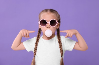 Photo of Girl in sunglasses blowing bubble gum on purple background
