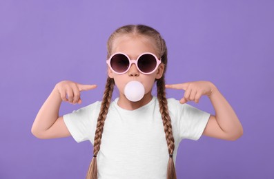 Girl in sunglasses blowing bubble gum on purple background