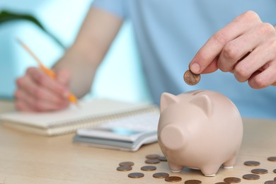 Financial savings. Man putting coin into piggy bank while writing down notes at wooden table, closeup