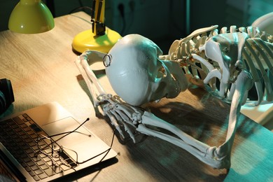 Waiting concept. Human skeleton sleeping at wooden table with laptop