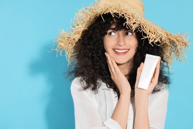 Photo of Beautiful young woman in straw hat holding tube of sun protection cream on light blue background, space for text