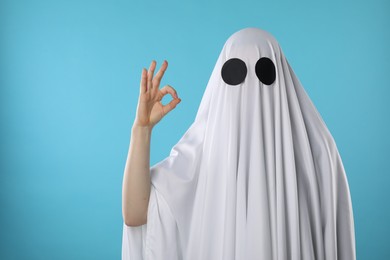 Photo of Funny ghost. Woman covered with white sheet showing OK gesture on light blue background