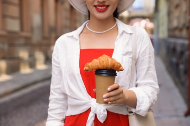 Photo of Woman holding tasty croissant and cup of coffee on city street, closeup