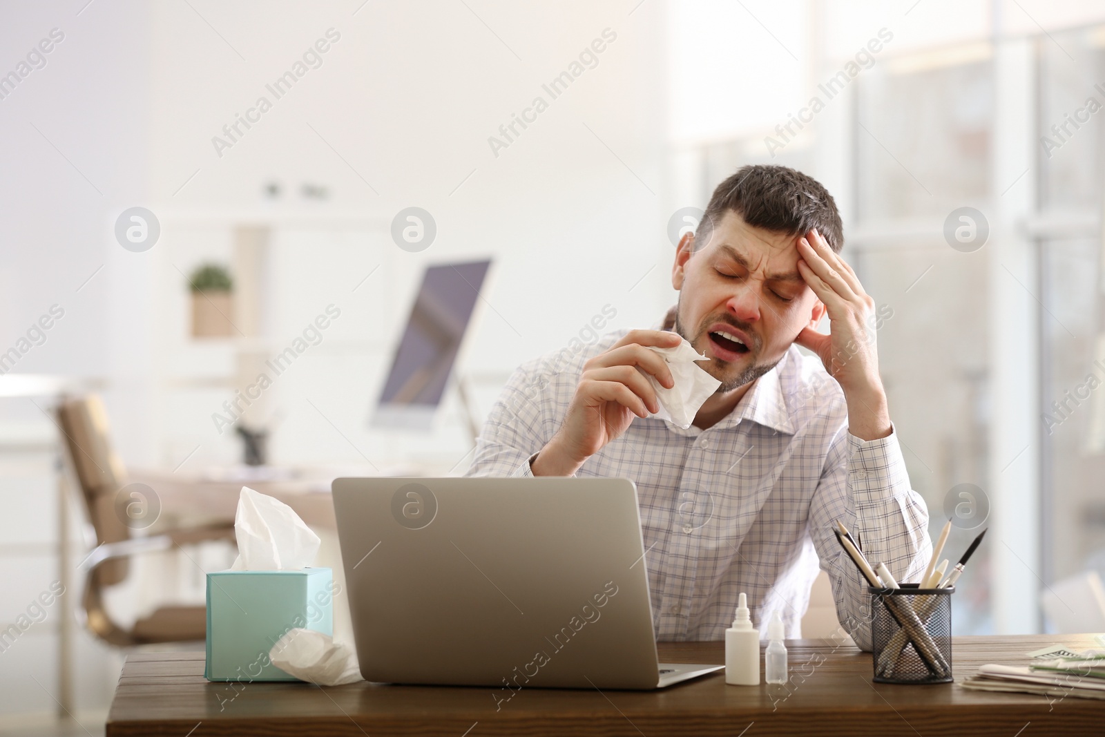 Photo of Ill man at table with nasal spray, drops and box of paper tissues in office