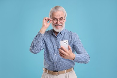 Portrait of stylish grandpa with glasses using smartphone on light blue background