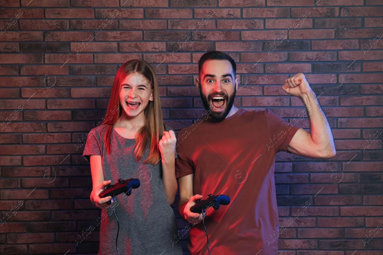 Photo of Young man and teenage girl playing video games with controllers near brick wall