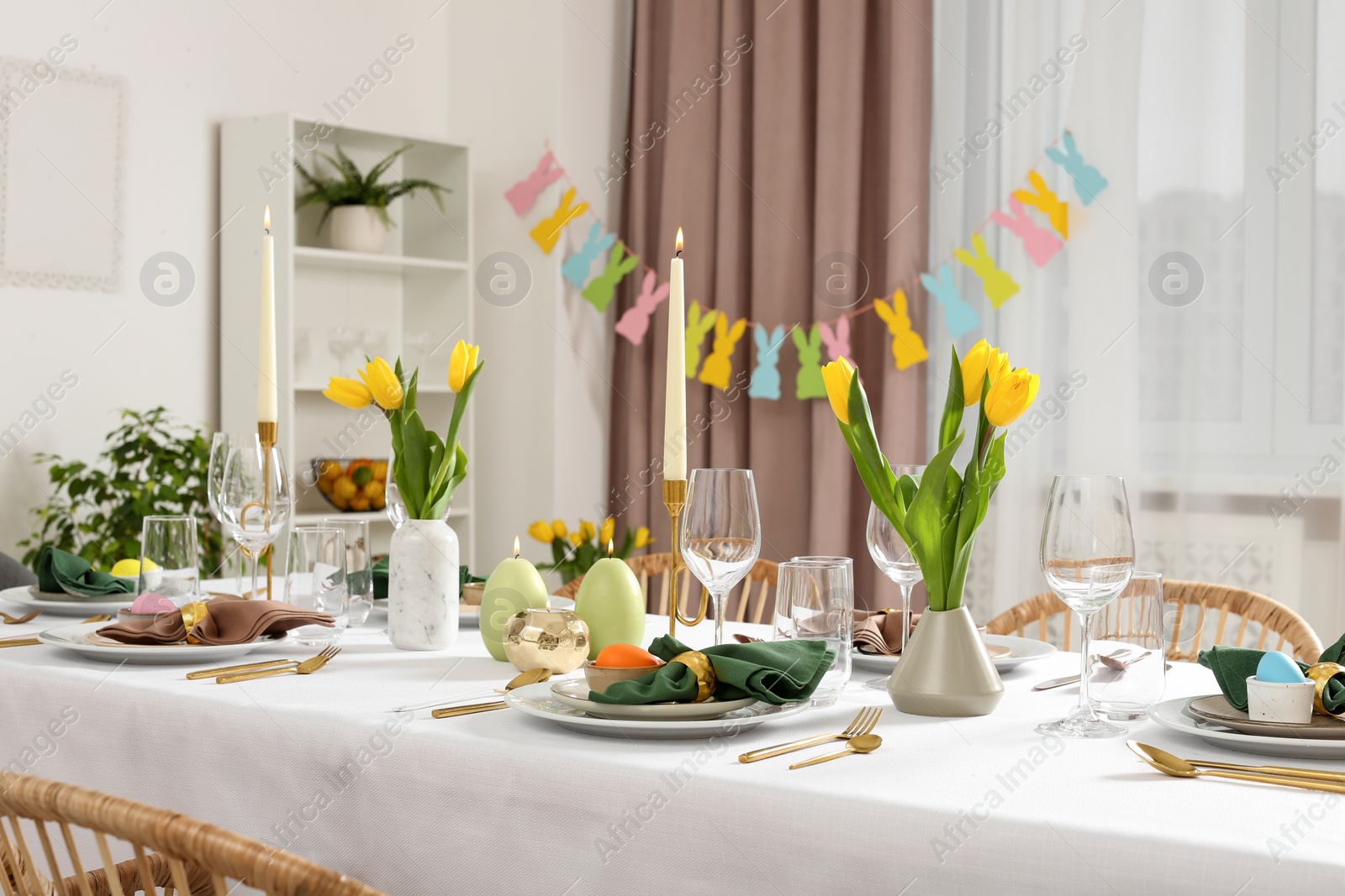 Photo of Festive Easter table setting with painted eggs, burning candles and yellow tulips in room