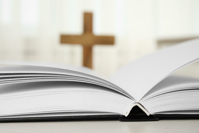 Photo of Open Bible on table and blurred cross on background, closeup. Christian religion