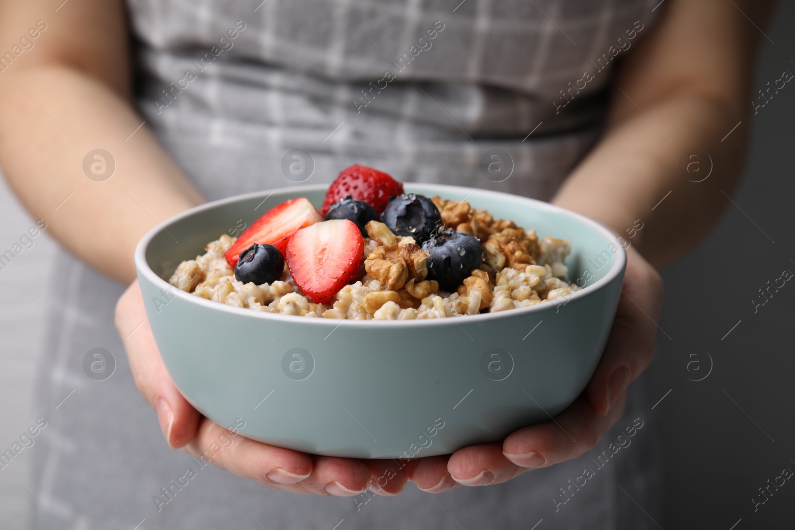 Photo of Woman holding bowl of tasty oatmeal with strawberries, blueberries and walnuts on grey background, closeup