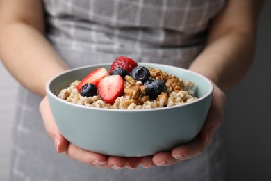 Photo of Woman holding bowl of tasty oatmeal with strawberries, blueberries and walnuts on grey background, closeup