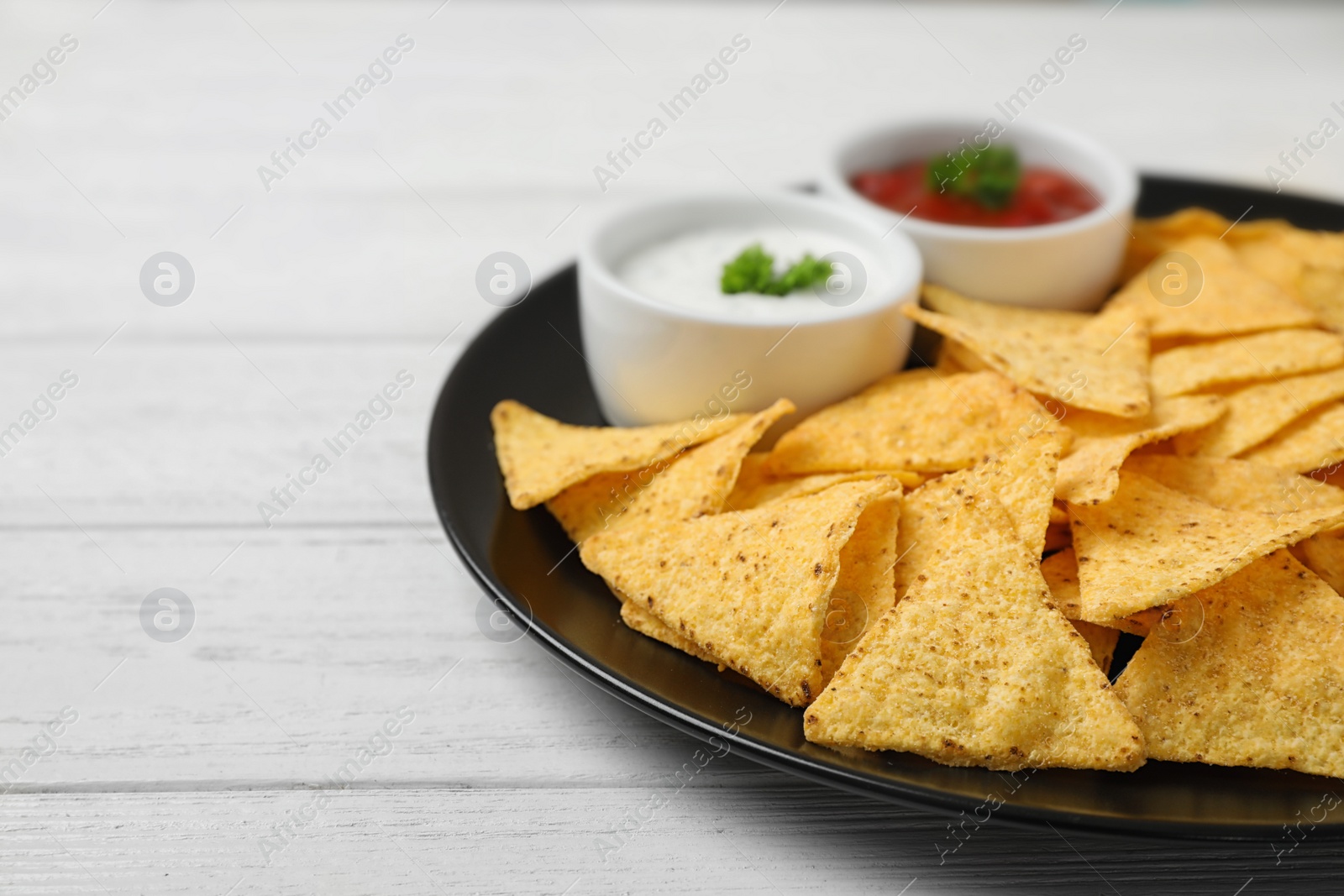 Photo of Black plate with tasty Mexican nachos chips and sauces on white wooden table, closeup. Space for text